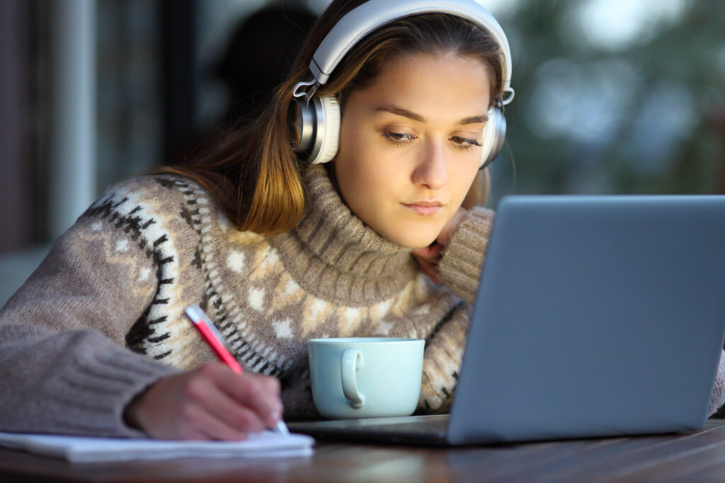 Concentrated student wearing headphones e-learning with laptop taking notes on notebook in a coffee shop in winter