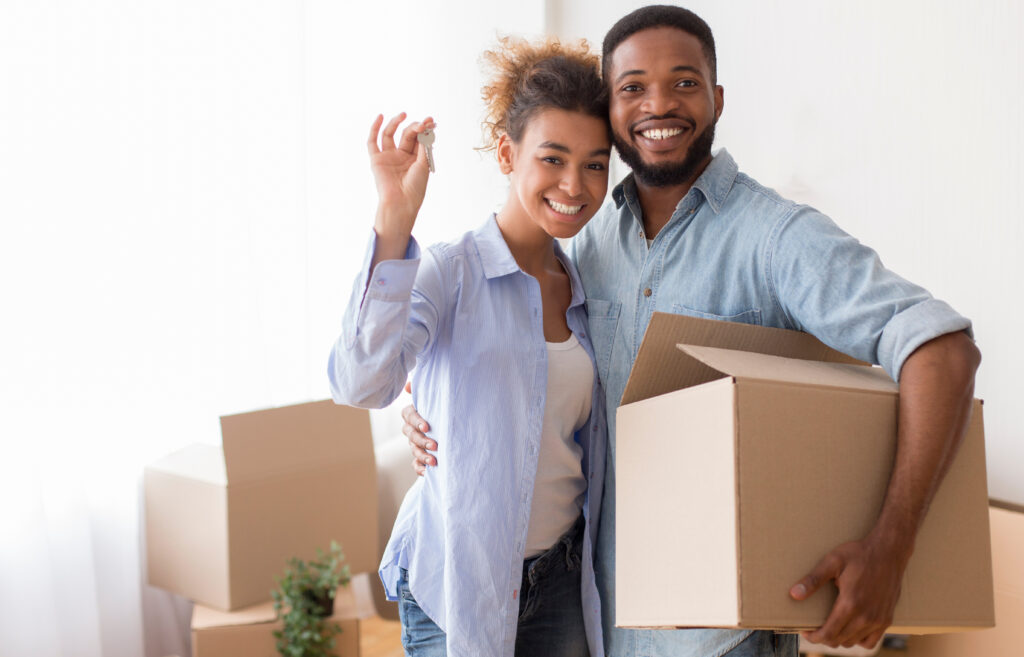 Happy Black Couple Showing Key Holding Moving Box And Hugging Standing In New Home.
