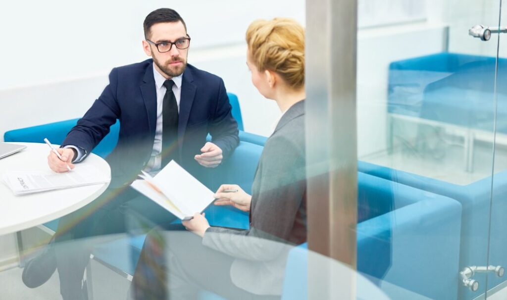 Two people in business attire sit across a round table in an office, having a discussion with documents and a notebook in hand.