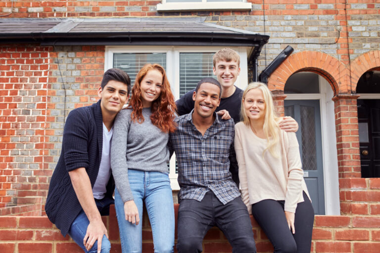 Five young adults smiling and posing together in front of a brick house